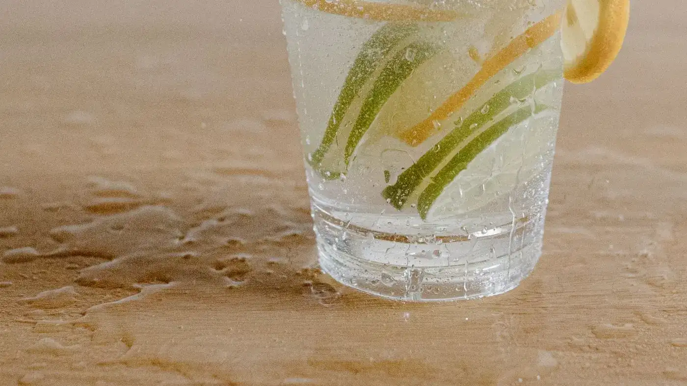 Vaso de vidrio con agua y rodajas de limón verde y amarillo sobre una mesa de madera con gotas de agua