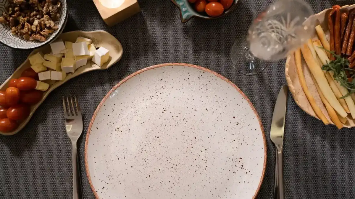Plato de cerámica vacío entre un tenedor y un cuchillo sobre un mantel gris, con tomates cherry, queso en cubos, nueces y palitos de pan alrededor