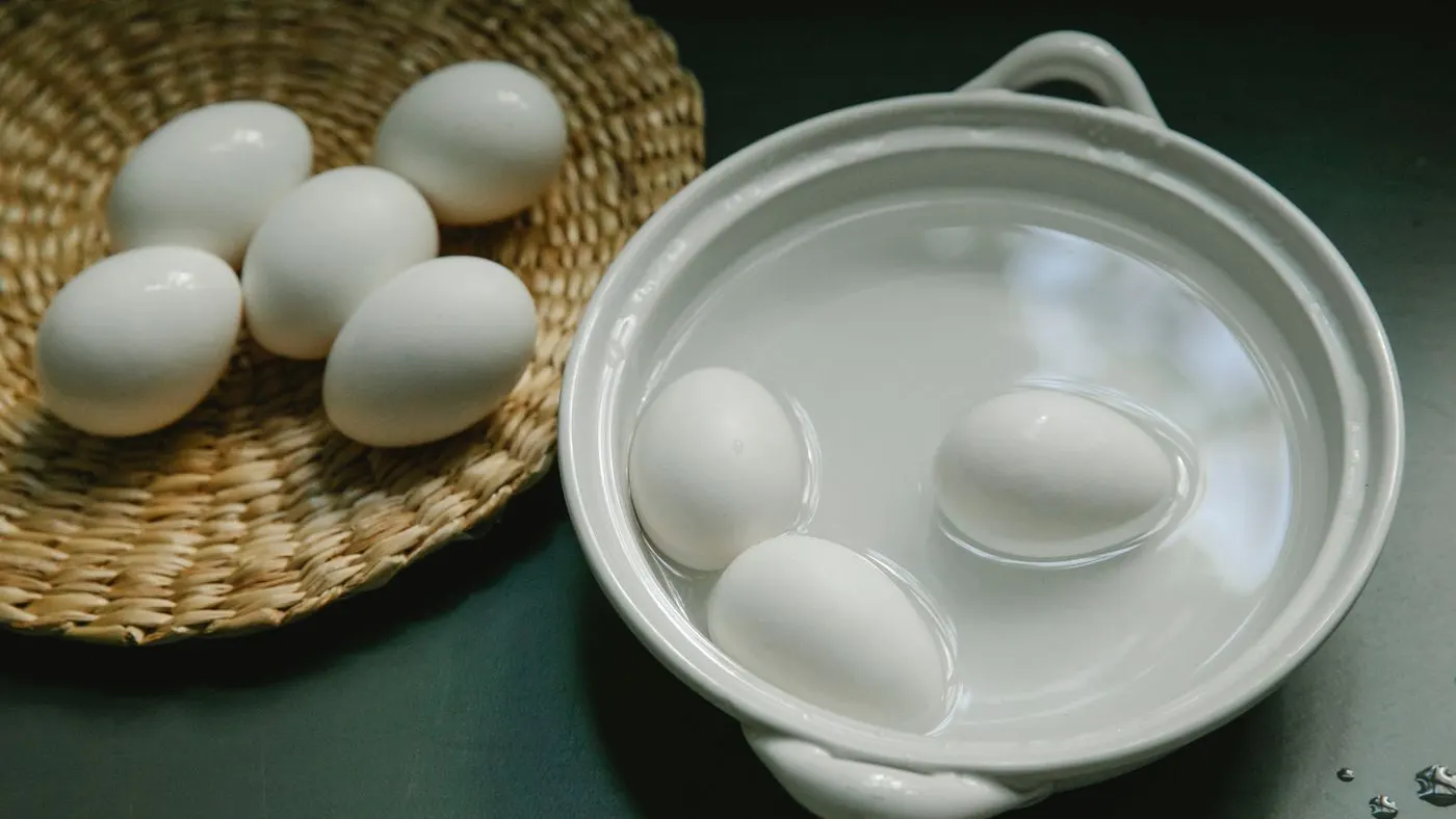 Huevos blancos cocidos en una olla de cerámica con agua y otros cinco huevos en un plato de fibra sobre una mesa oscura, para contar las calorías de un huevo