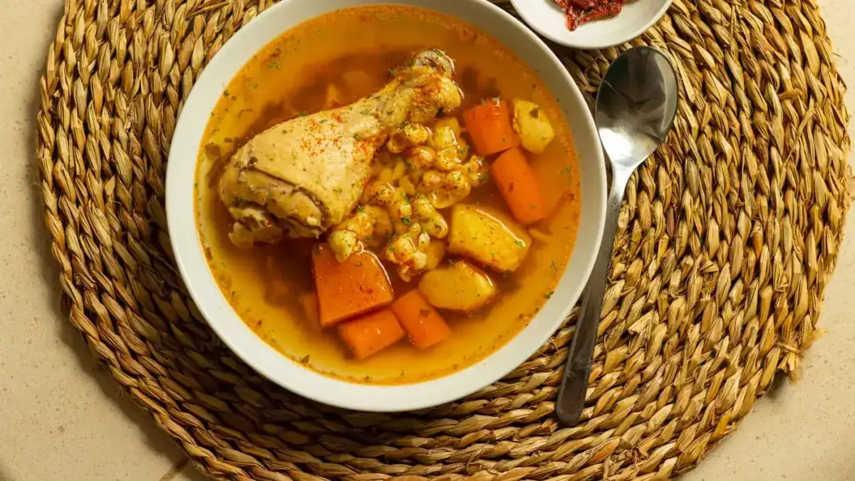 Sopa de pollo con papa y zanahoria en un tazón blanco sobre un individual de fibra, con una cuchara al lado