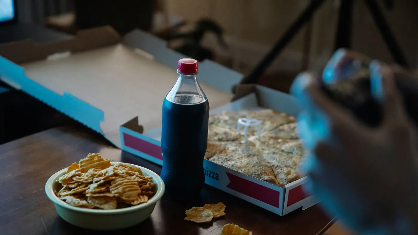 Mesa de madera con un tazón de papas fritas de paquete, una botella de gaseosa oscura y una caja de pizza abierta