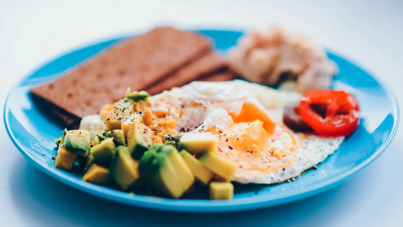 Plato azul con un huevo frito, aguacate picado, pimentón rojo y tostadas oscuras: un desayuno casero ordenado