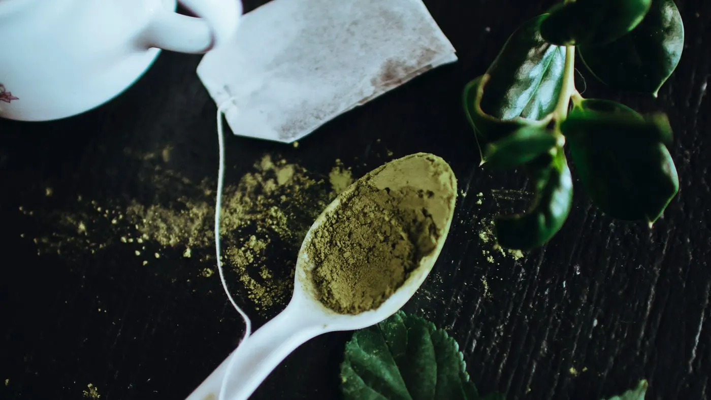 Cuchara blanca con polvo verde de té sobre una mesa oscura, entre hojas verdes