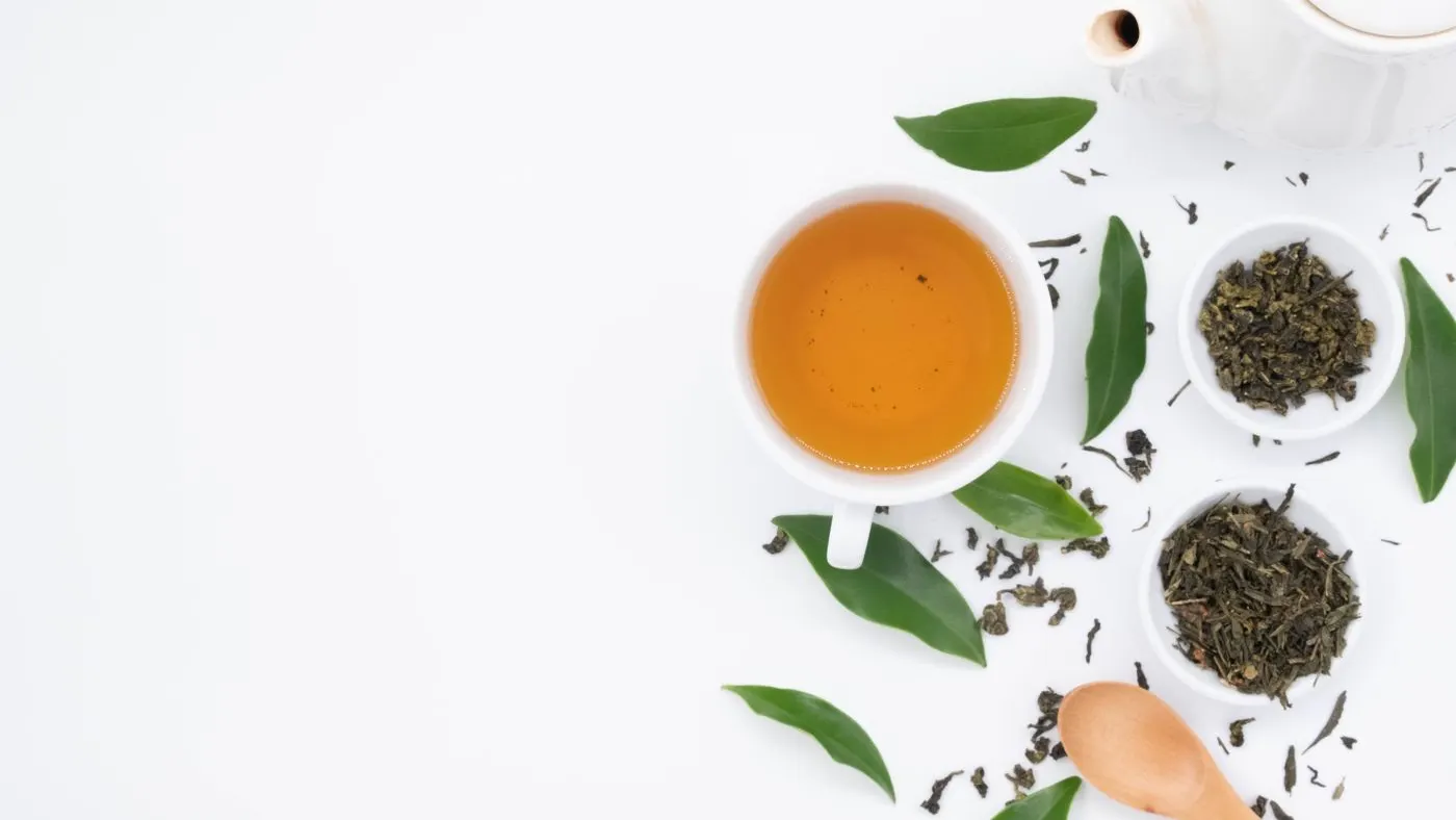 Taza blanca con té verde, hojas secas en dos platillos, una cuchara de madera y una tetera blanca sobre fondo claro